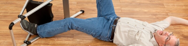 An elder man has fallen off of a stool and is laying on the floor.